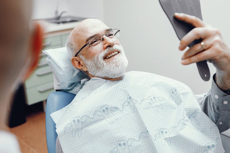 An image of an older man sitting in a dentist chair and smiling after getting his new dental implants from our implant specialist in Voorhees, NJ.